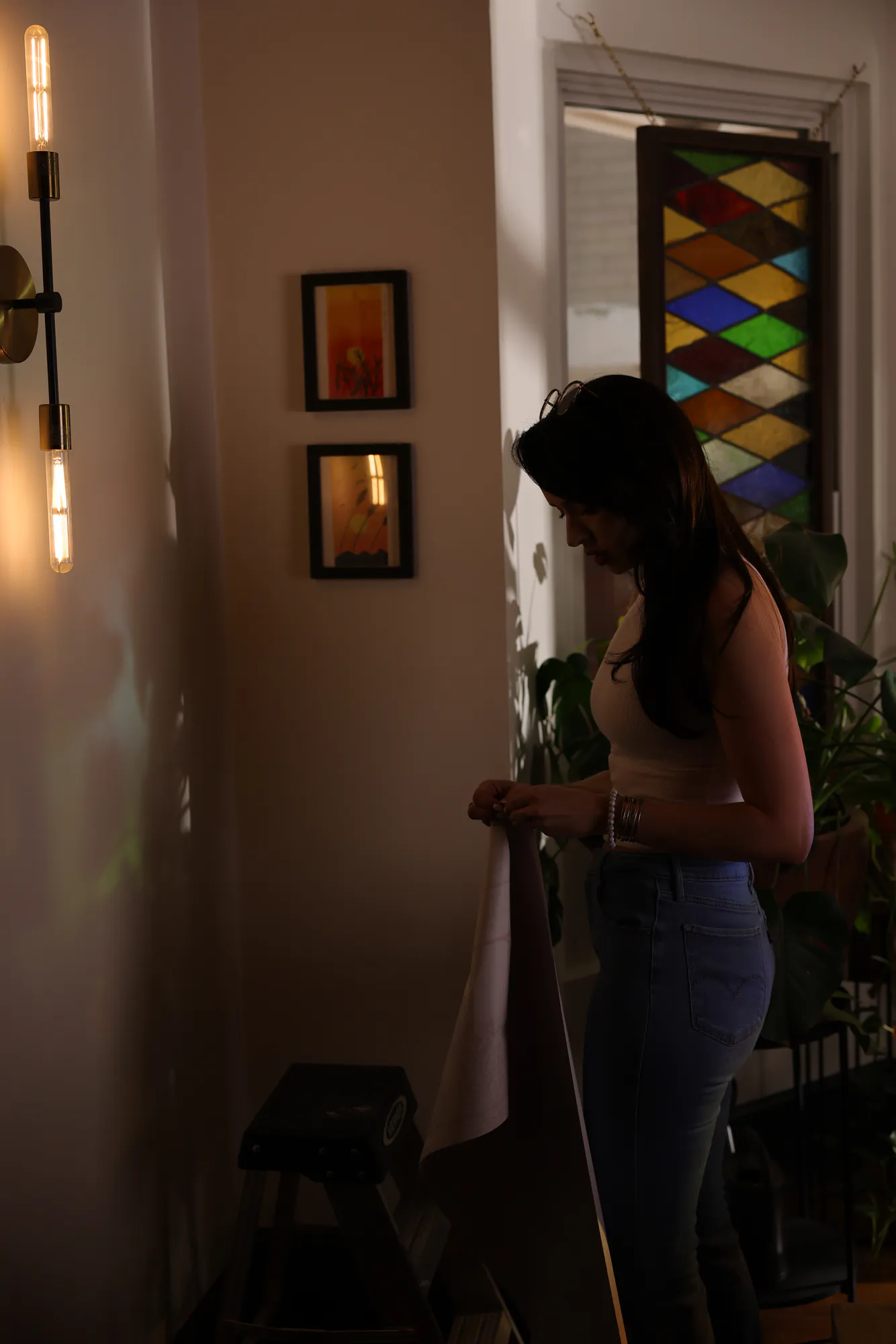 Leticia Marie Holtz holding a length of wallpaper against a hallway wall in low evening light, checking the colour beside a brass bulb sconce, two small framed prints and a leaded stained-glass window