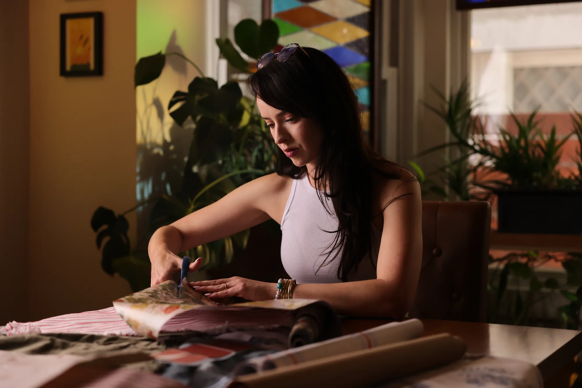 Leticia Marie Holtz seated at a dining table cutting into a fabric swatch book, rolls of wallpaper and textile samples stacked beside her, monstera leaves and a diamond-pane stained-glass window behind