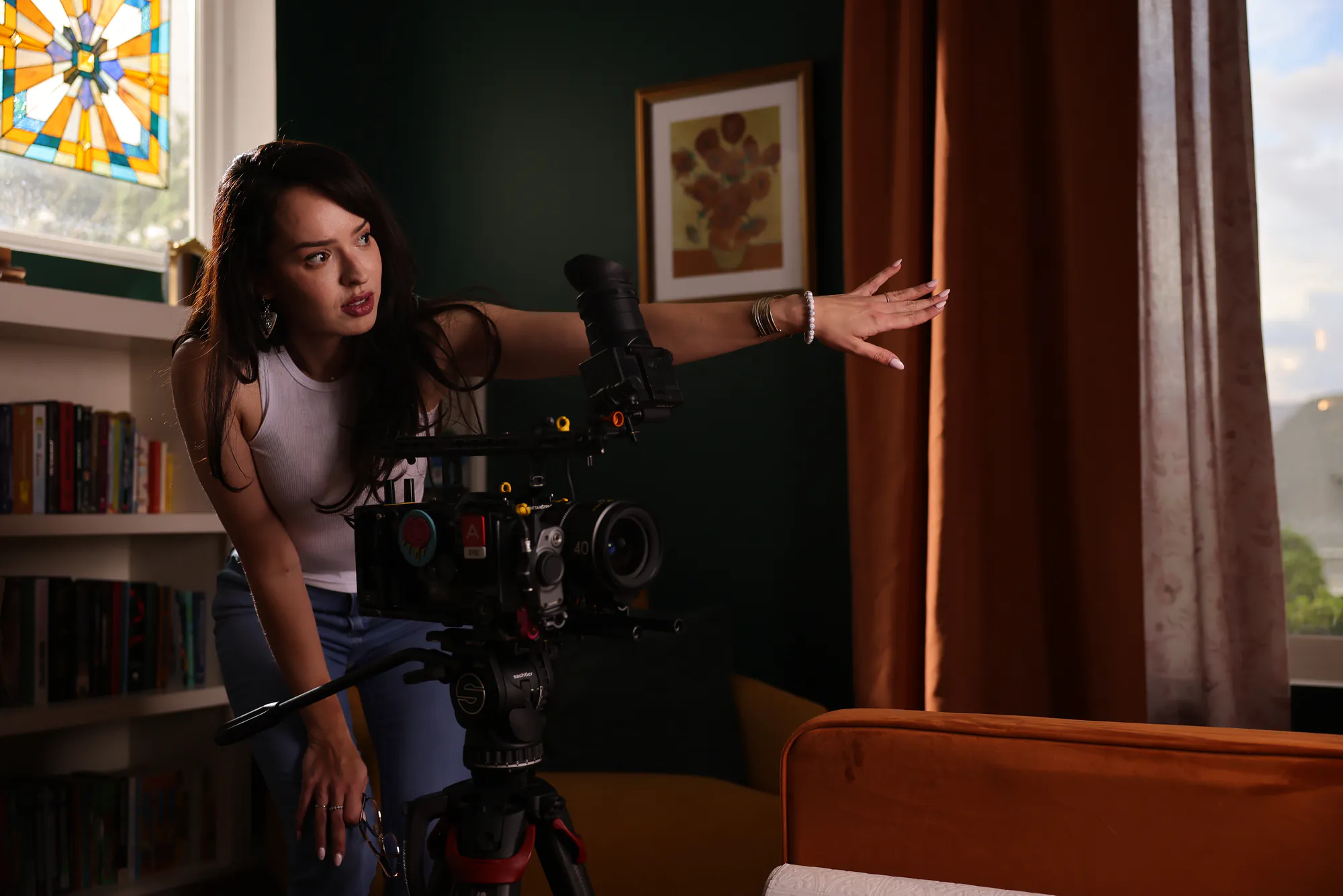 Leticia Marie Holtz leaning over a cinema camera on a tripod, one arm extended to block a shot across a dressed living room with deep green walls, a stained-glass window, a framed sunflower print and rust velvet curtains